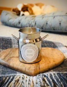A Milk churn shaped candle by The Cattleshed Candle Company on a heart shaped wooden board displayed on a blanket