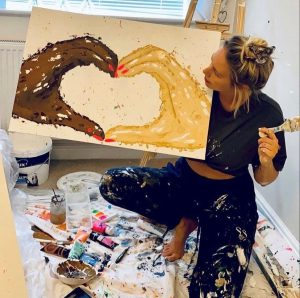 Artist Cherylee Scarsbrook sat on the floor holding one of artworks of two hands, one black, one white, coming together to form a heart shape with their fingers