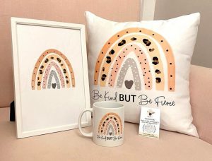  A mug, cushion and framed print all showing the same illustration of a pink, grey and black rainbow displayed on a table
