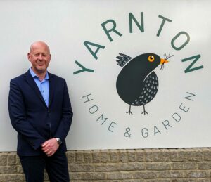 Image of a man in a navy suit and sky blue shirt posing in front of the logo Yarnton Home & Garden