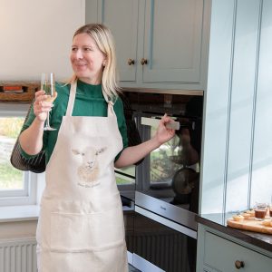 A lady (Liz Corely_ with a glass of white wine in her hand and wearing an apron in her kitchen