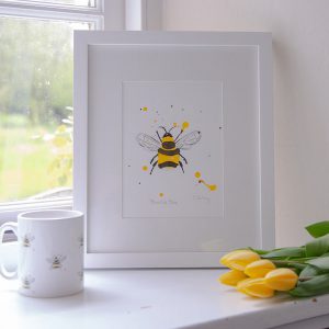 A framed picture of a hand-painted bee on a window sill next to a bee printed mug and some bright yellow tulips.. Mug and iframed illustration by Liz Corley Art.