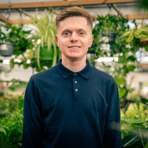 A clean-shaven young man with brown short hair wearing a navy blue polo top button to the neck and standing in front of a bright and green display of house plants