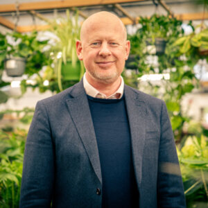 A head and shoulders photo of a man in a navy jacket and jumper and white shirt, with lush green house plants displayed in the background.