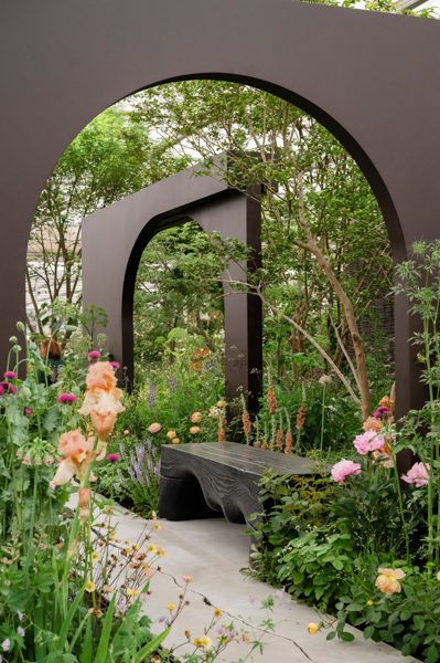 A grey arch framing a garden bench and spiralling pink and peach flowered garden plants