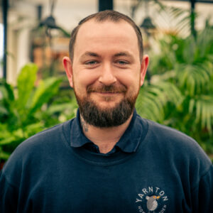A head and shoulders shot of a man with brown hair and a beard dressed in a dark blue shirt and a sweatshirt which has a company logo on it Yarnton Home & Garden