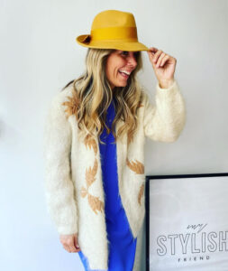 A woman with long blonde hair wearing a bright yellow trilby. She is laughing and tipping the hat slightly. she is wearing a brilliant blue jumpsuit and a long cream cardigan.