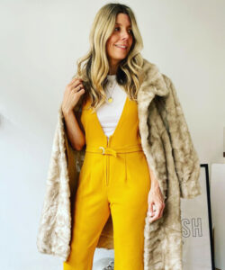 A tall woman with long blonde hair wearing a white shirt and bright yellow jumpsuit. A faux fur beige coat is draped over her shoulders.