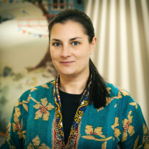 A lady wearing a turquoise kimono style top with yellow flowers. Her dark brown hair is swept off her face in to a ponytail. She has brown eyes and a natural complexion.